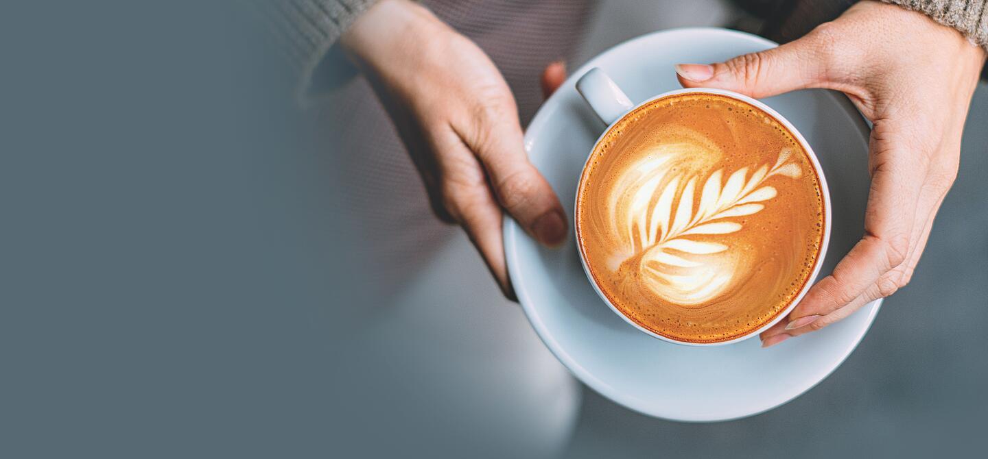 Zwei Hände halten Dallmayr Cappuccino mit Latte Art Blatt