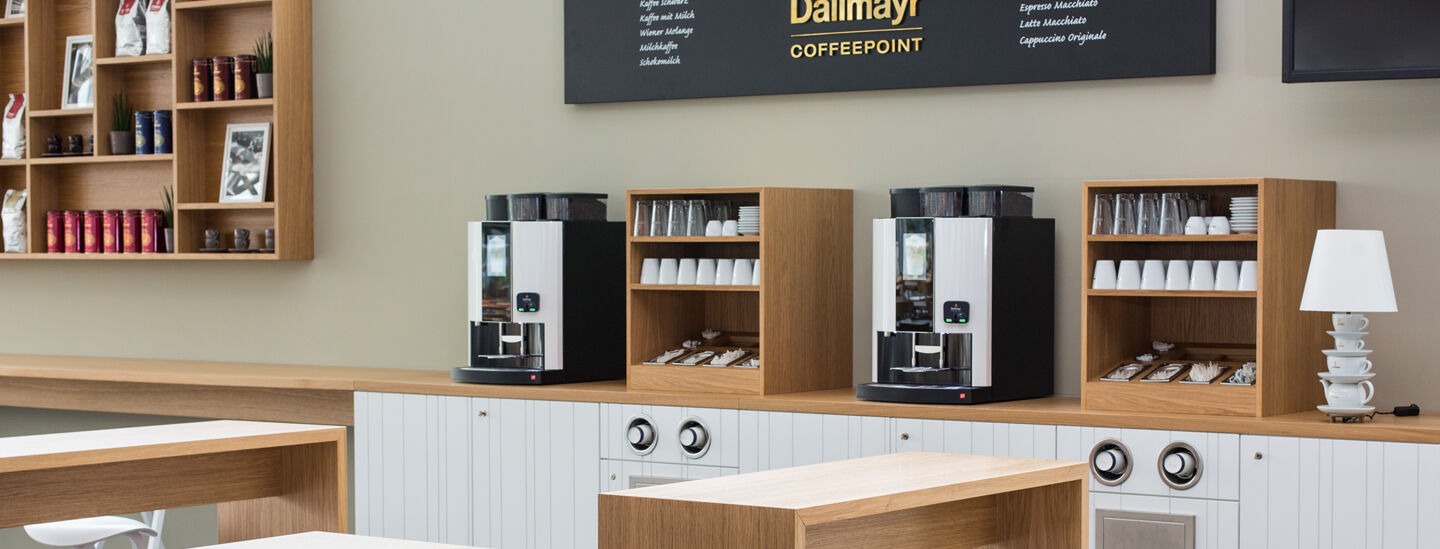 Plnoautomatické kávovary Dallmayr na príborníku so šálkami a príslušenstvom