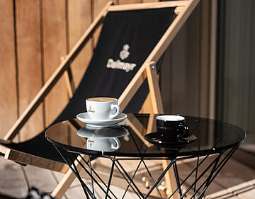 Cappuccino și espresso Dallmayr în căni, pe o măsuță laterală de lângă un șezlong Dallmayr