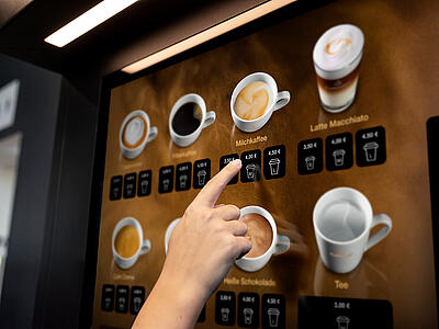 Un client sélectionne un produit à base de café sur l'écran numérique d'une machine automatique Dallmayr.