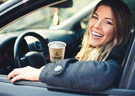 A woman drinking from a Dallmayr coffee to-go cup in a car at a petrol station
