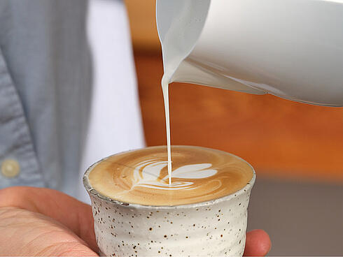 A barista creating latte art in a grey Dallmayr cup